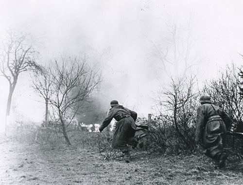 Two German soldiers advance towards burning vehicles